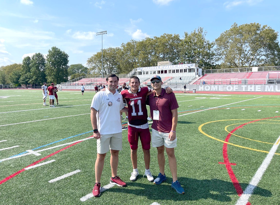 Three young men, the middle in a football uniform, stand on a football field and smile at the camera