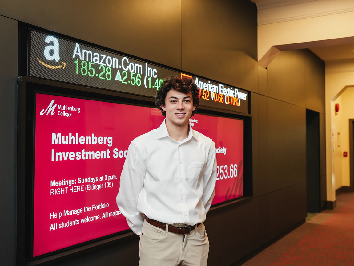 A college student in a white button down and khakis stands in front of a stock ticker