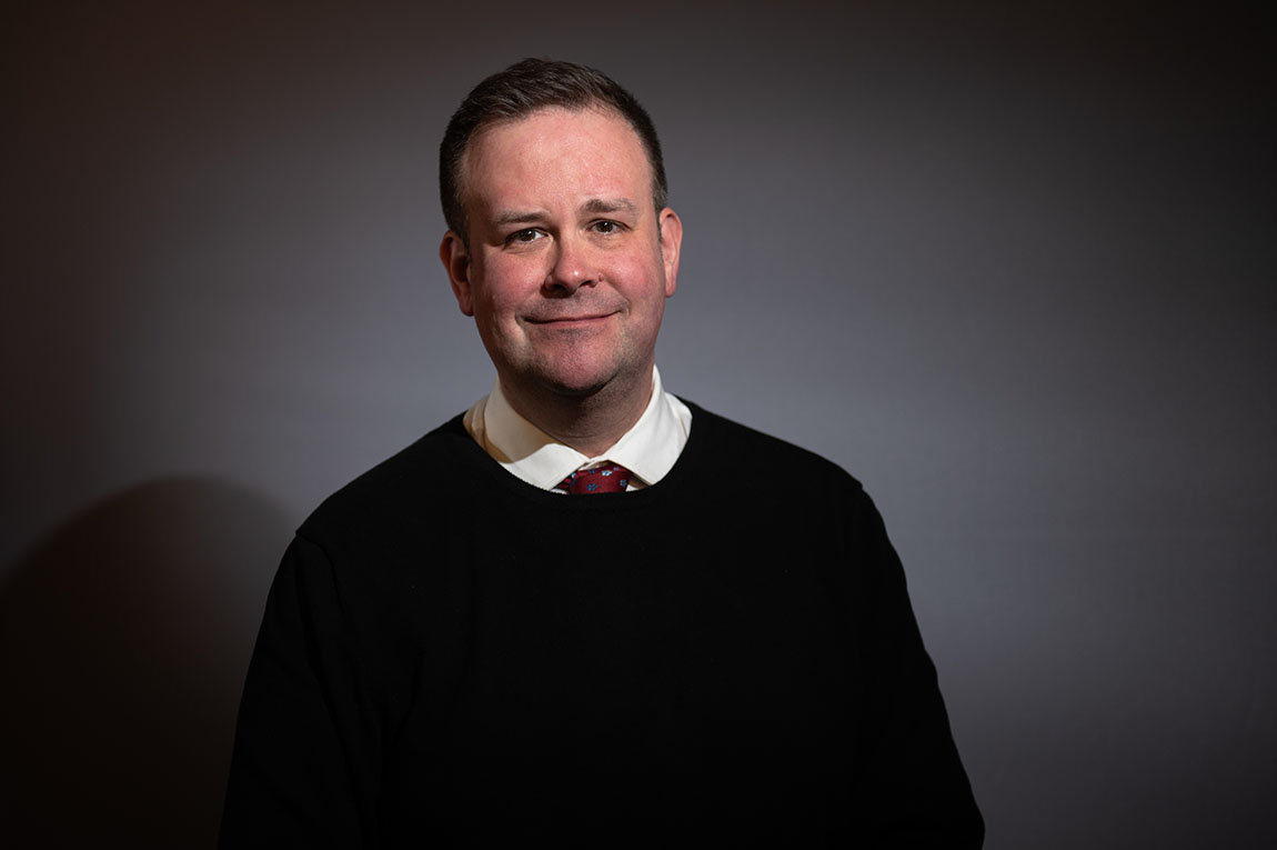 A white male with brown hair and necktie poses for a studio portrait