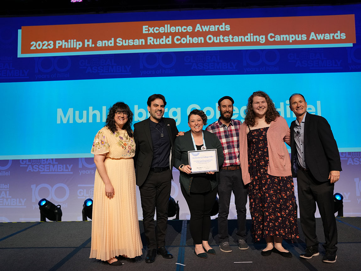 Six people stand on stage smiling with the person in the center holding a certificate