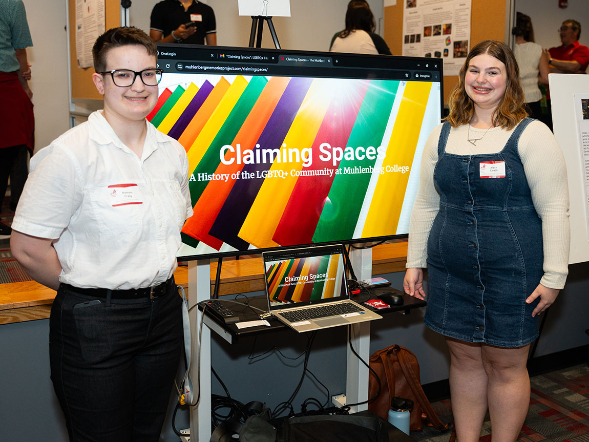 Two college students stand in front of a video screen presentation; text on the screen says Claiming Spaces