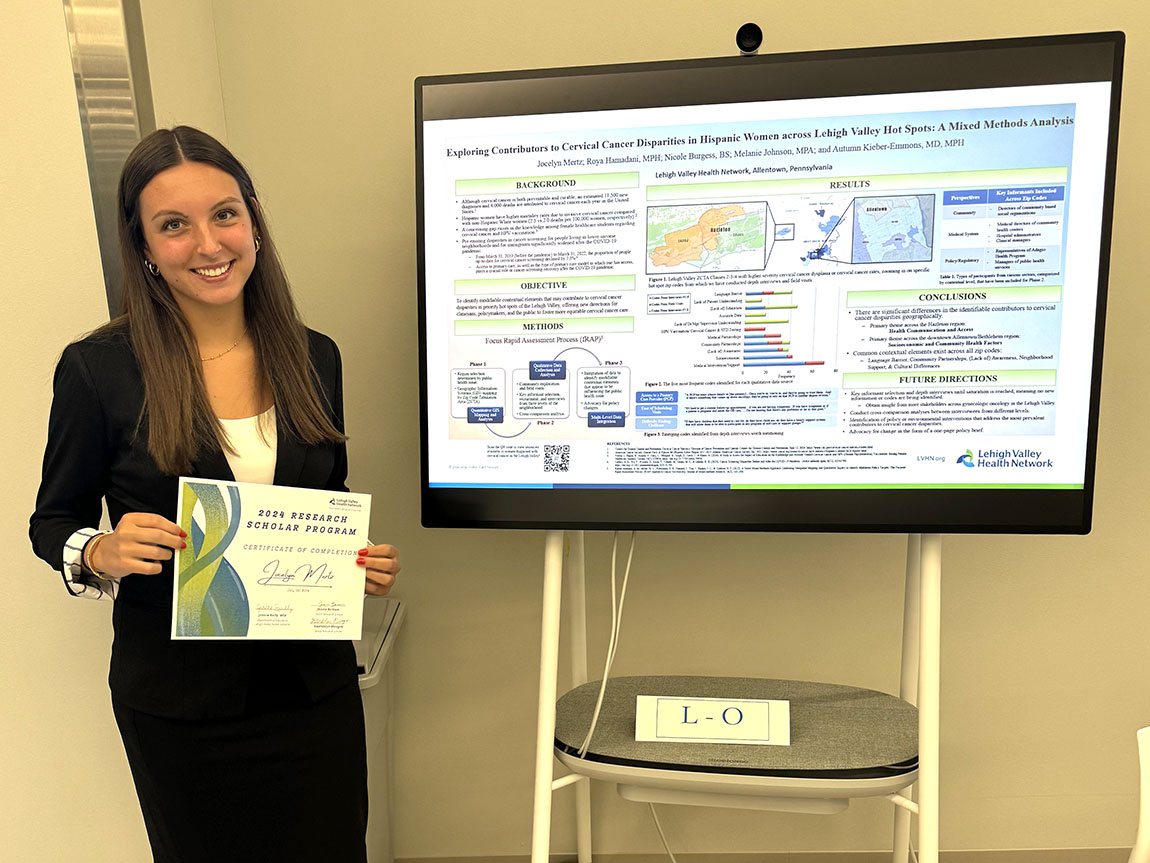 A college student smiles while holding a certificate next to an academic poster