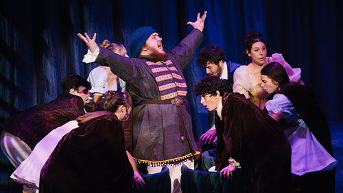 Actor in ornate costume stands center stage with arms raised as ensemble surrounds him in a dramatic scene.