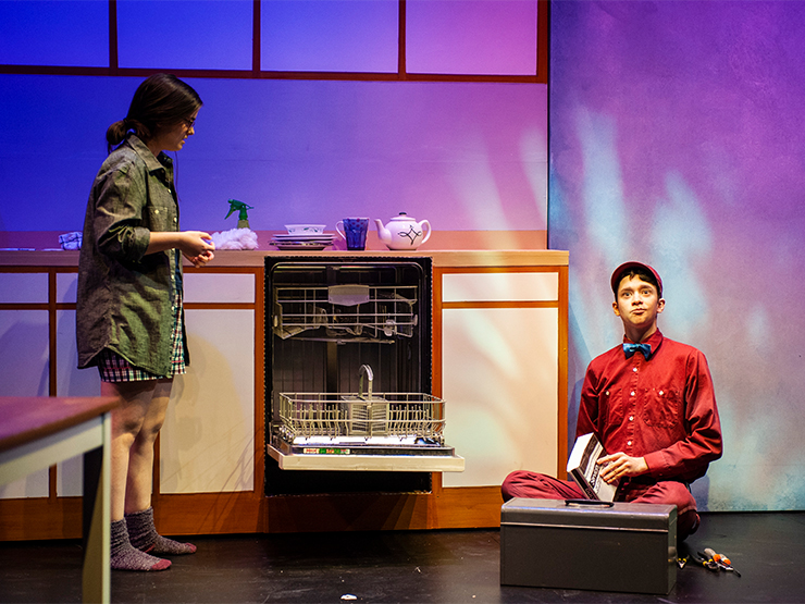 A student stands on one side of an open dishwasher and a student, dressed as a repair man, sits on the floor on the other side.