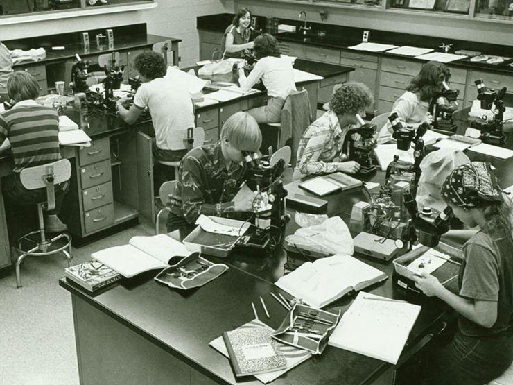 A black and white image of a lab full of students, dressed in clothing from the 1970s, working with microscopes.