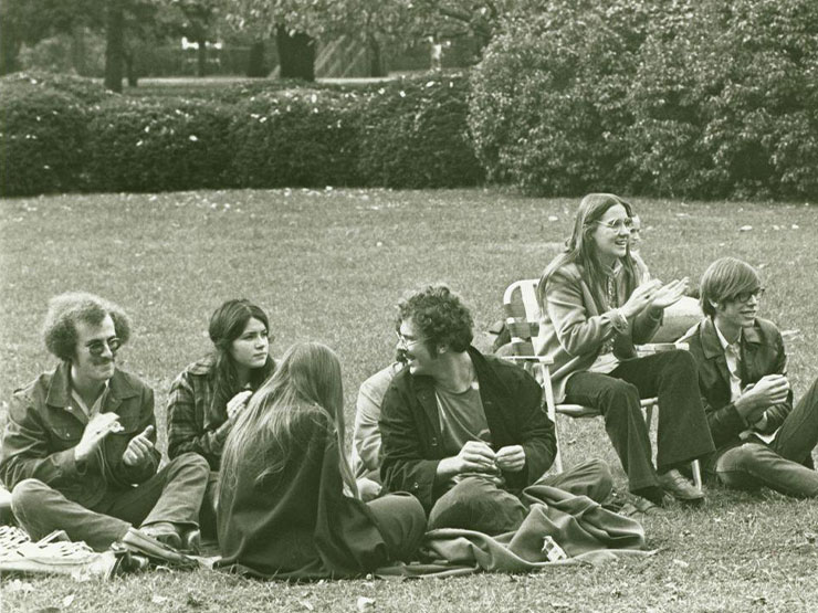 A black and white image of a group of college students dressed in clothing from the 1970s, sitting together on the grass.