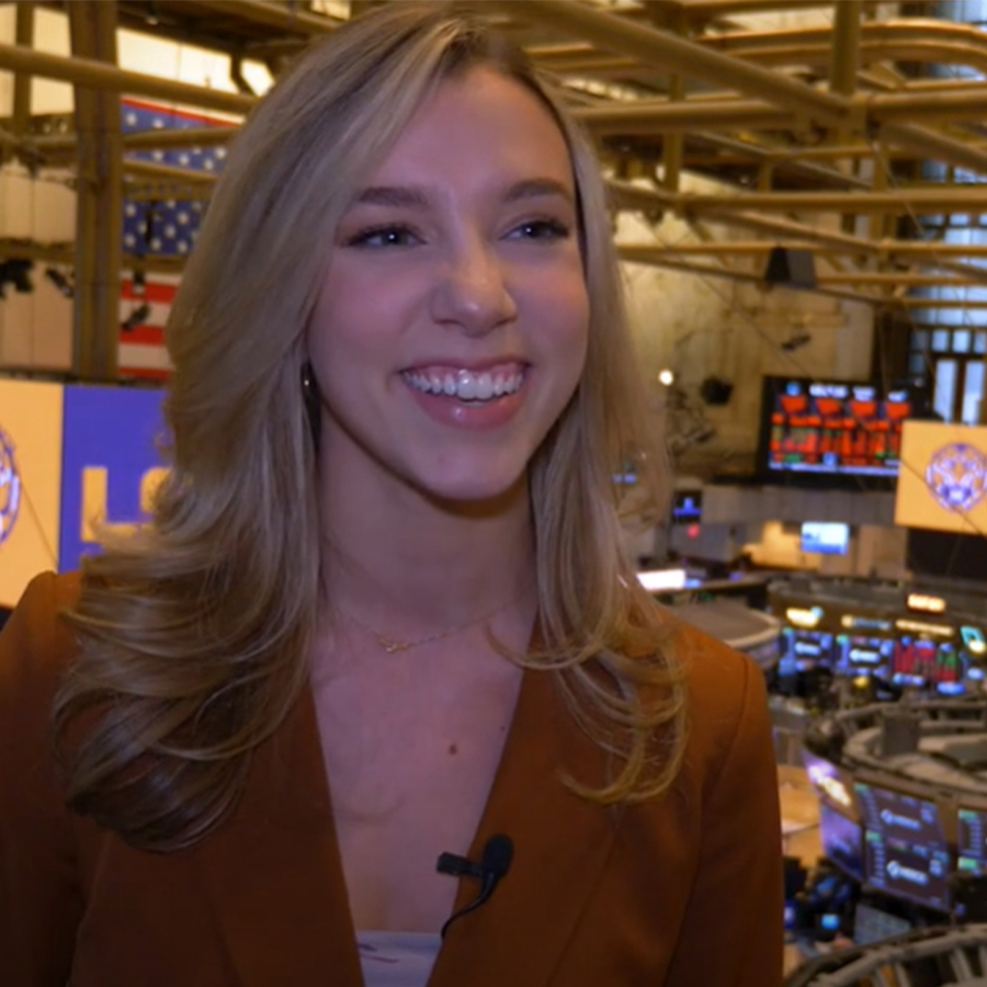 A woman dressed professionally speaks the camera from the floor of the New York Stock Exchange.